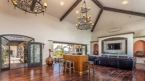A spacious living area with a wooden floor, a bar with stools, a large black leather sofa, and a television mounted on the wall.