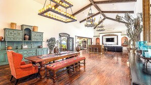 A spacious living room with a large wooden dining table, a red armchair, and a bench. There are multiple hanging chandeliers and a large TV on the wall.