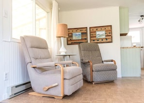 Twee grijze fauteuils met houten leuningen, een witte vloerlamp, en twee kunstwerken met houten lijst aan de muur.