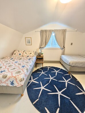 A bedroom with two beds, a window with curtains, a bedside table with a lamp, and a star-patterned rug.