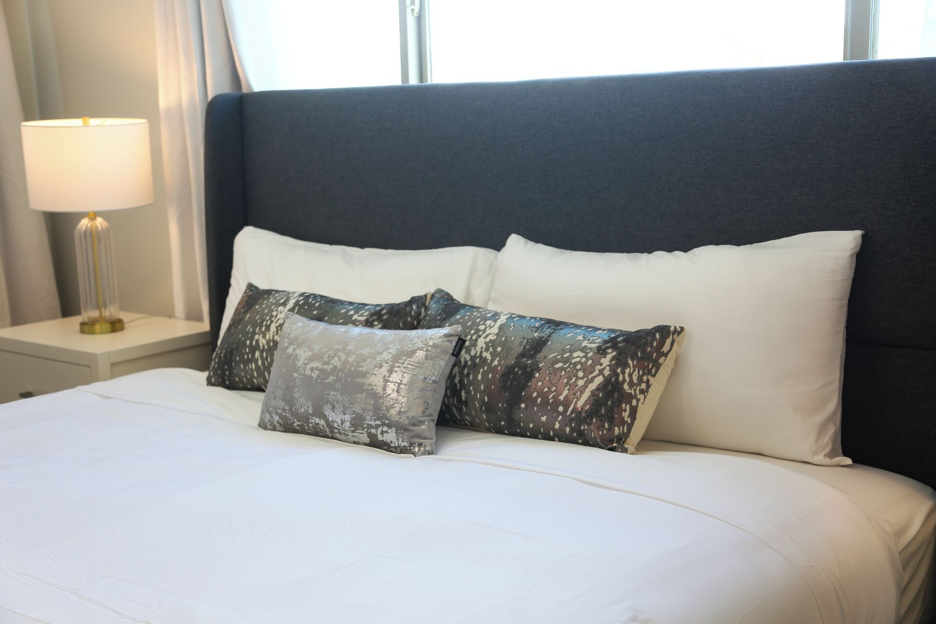 A neatly made bed with white linens and two patterned pillows, a bedside lamp, and a dark headboard.