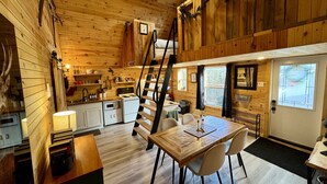 A cozy cabin interior with a wooden interior, a dining area with a table and chairs, a kitchen with a stove and cabinets, and a loft bedroom with a ladder.