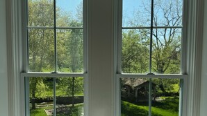 Um quarto com duas janelas grandes, cada uma dividida em quatro painéis, com vista para um jardim bem cuidado e um edifício ao longe.