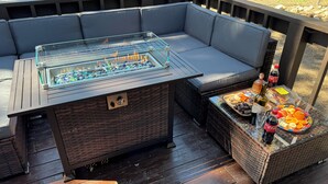 Ein Außenbereich mit Sitzgelegenheit, einem Tisch mit Glasplatte, einem eingebauten Grill und einem Korbtisch mit Snacks und Getränken.