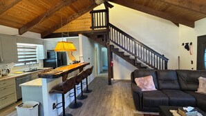 Eine geräumige Küche mit einer zentralen Kochinsel, ein Essbereich mit Barhocker sowie eine schwarze Ledersofa und eine Holztreppe.