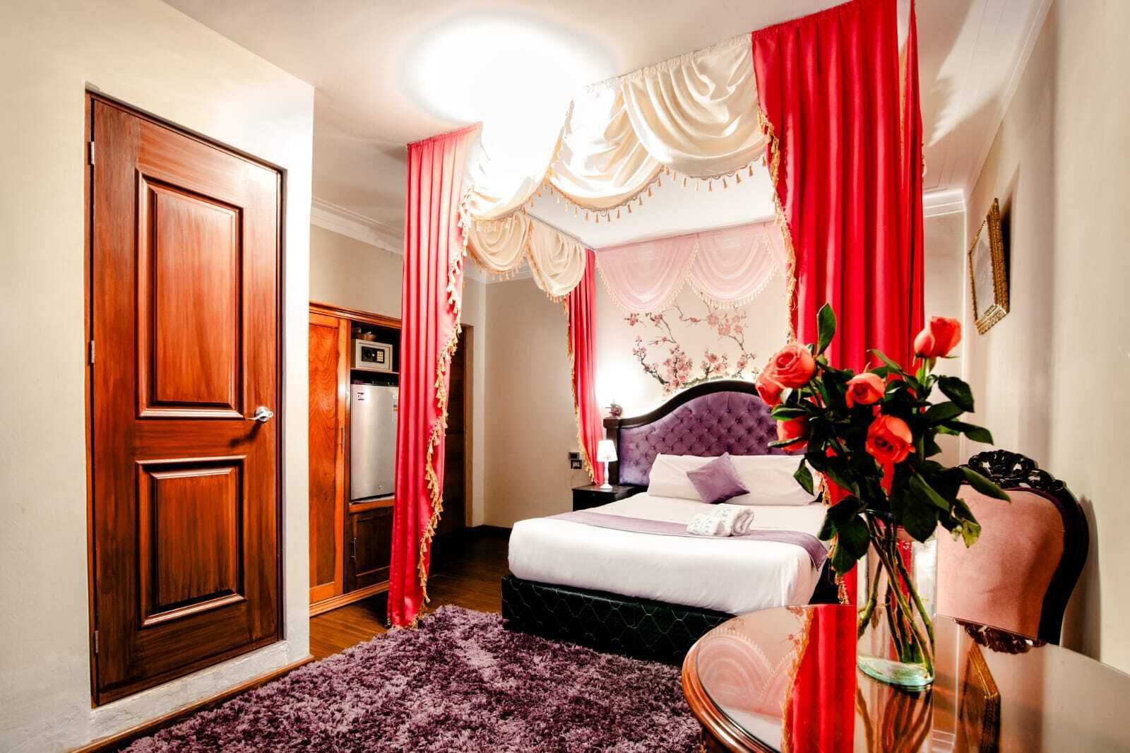 A bedroom with a wooden door, a bed with a purple headboard, a red and white curtain, and a vase with flowers.