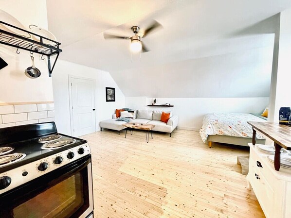 A compact kitchen with a stove, a ceiling fan, a sofa, and a bed in a small room with wooden flooring. - Finn`s Studio Suite Apartment, Alma, Fundy Park (Alma)