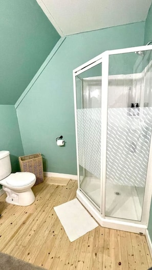 Un baño con inodoro blanco, una cesta de mimbre, cabina de ducha blanca y un tapete en el suelo.