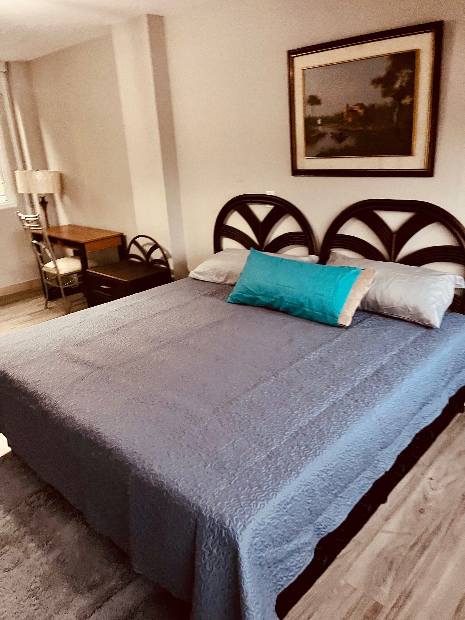 Un letto rifatto con una coperta grigia e due cuscini, un comodino in legno con una lampada e un quadro appeso al muro.