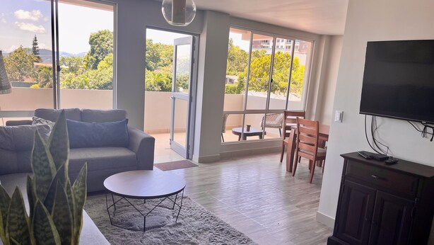 Una sala de estar moderna con un sofá, una mesa de centro redonda, un televisor y un balcón con mobiliario exterior.