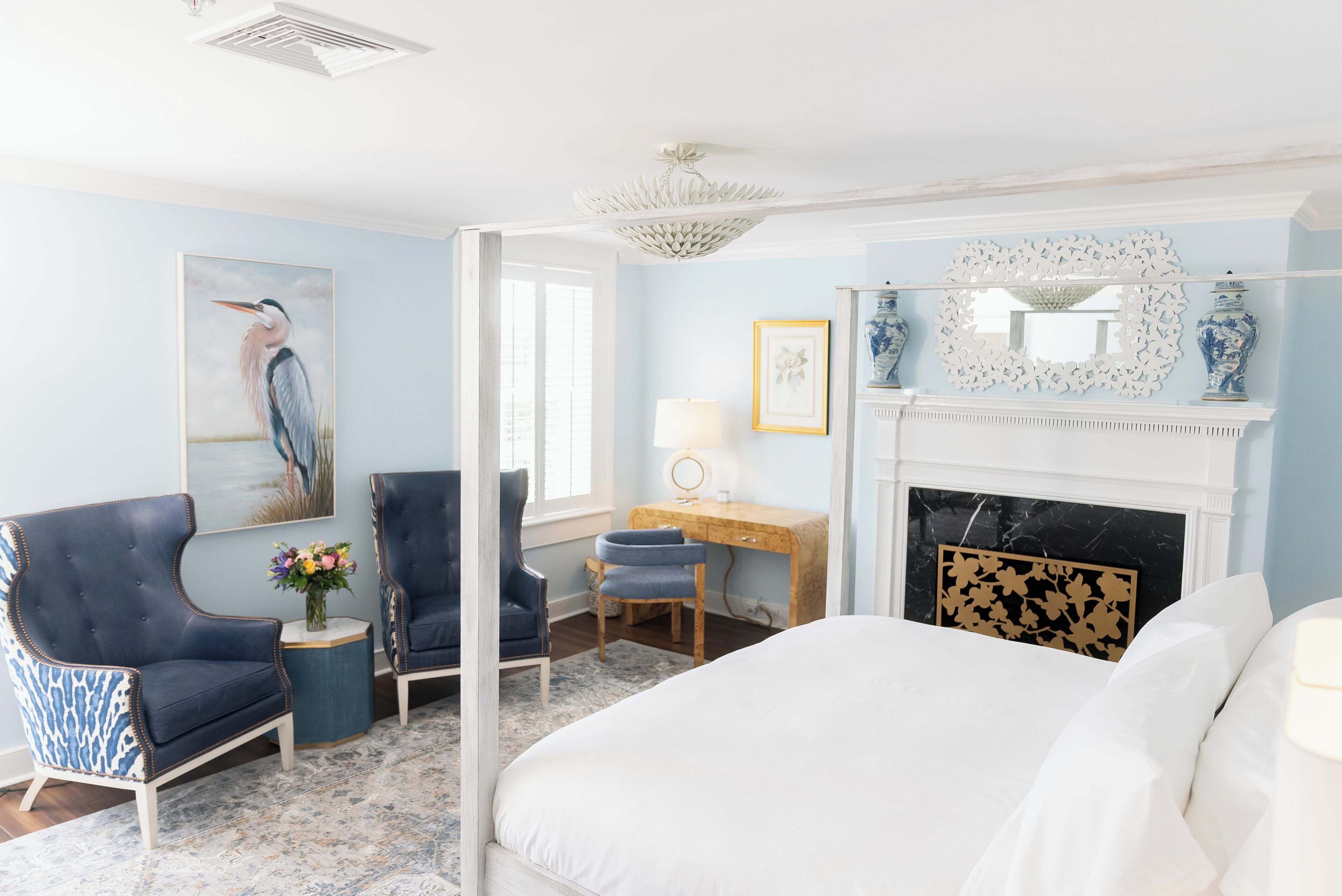 A bedroom with a bed, two armchairs, a desk, a fireplace, and a painting of a bird.