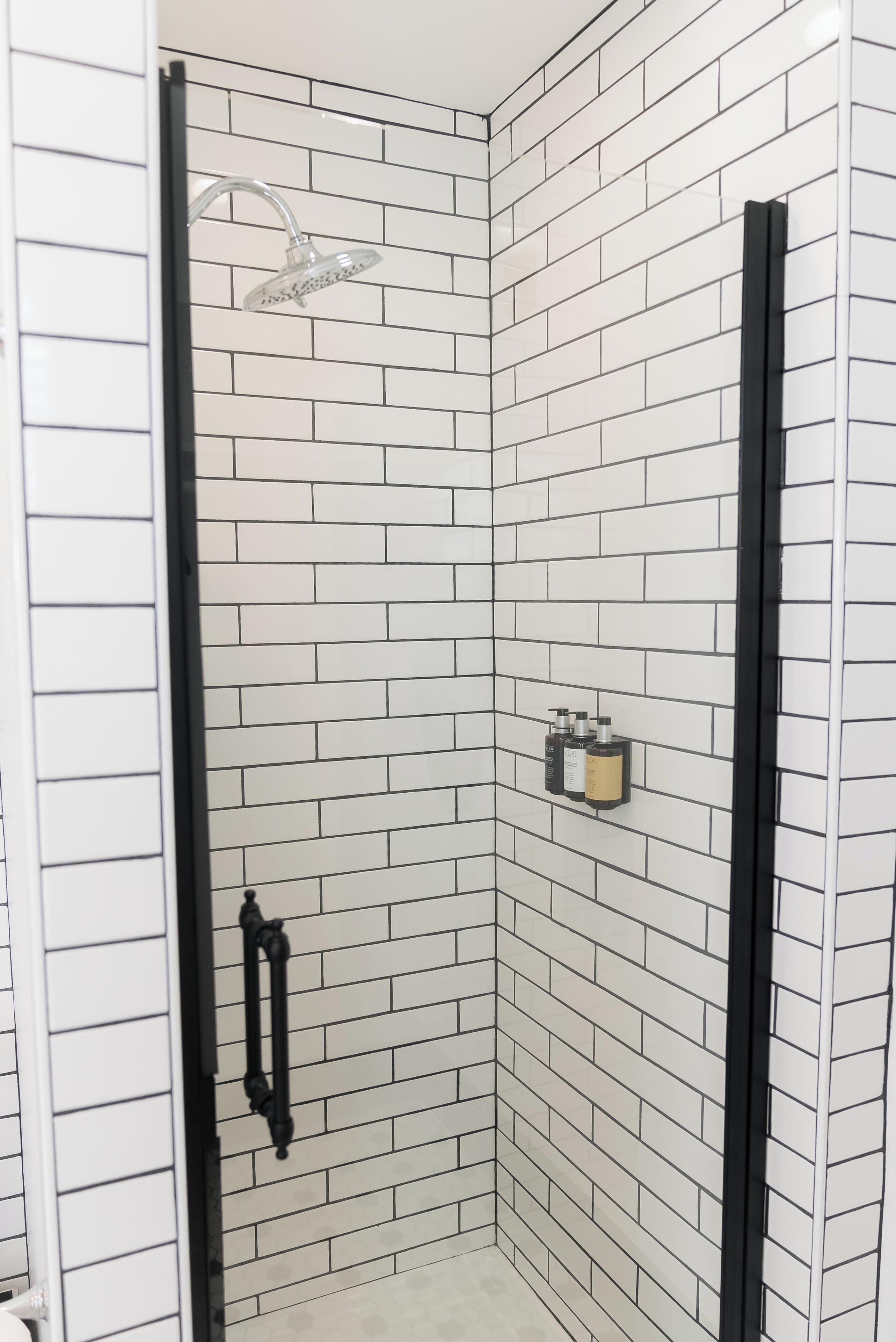 Um chuveiro no canto, com paredes revestidas de azulejos brancos e moldura de porta preta.