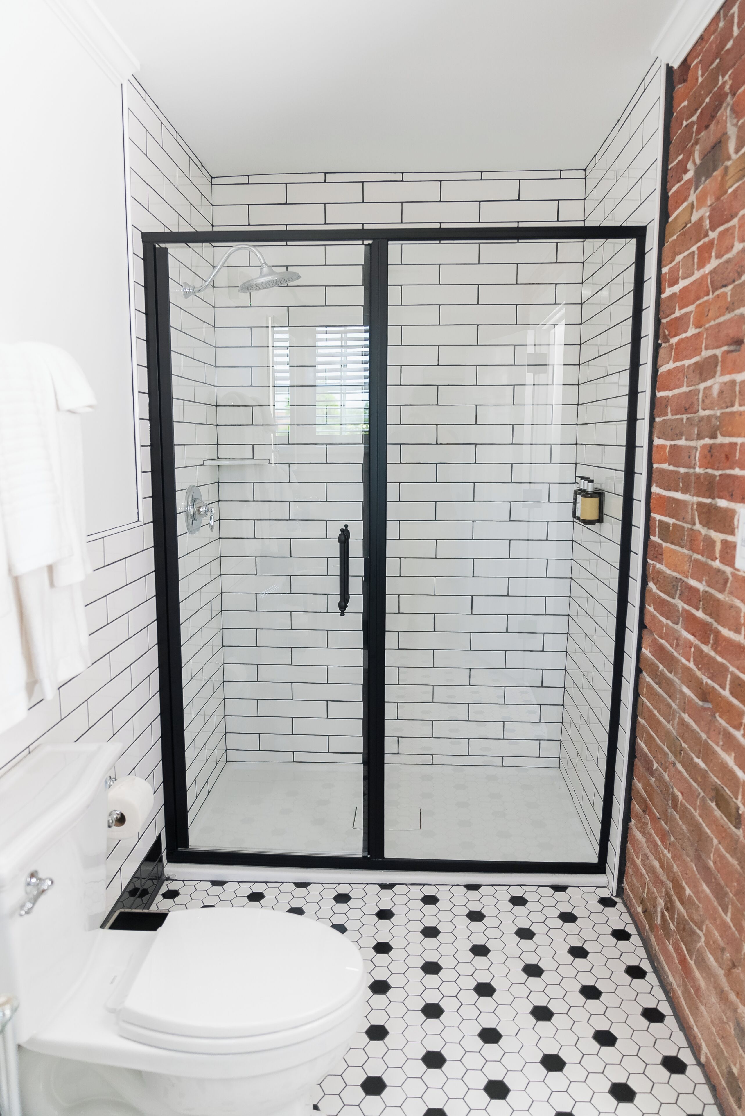 A bathroom with a glass-enclosed shower, white tiled walls, a black-framed shower door, a white toilet, and a black and white hexagonal tile floor.