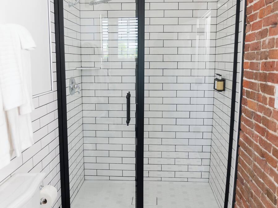 A bathroom with a glass-enclosed shower, white tiled walls, a black-framed shower door, a white toilet, and a black and white hexagonal tile floor.