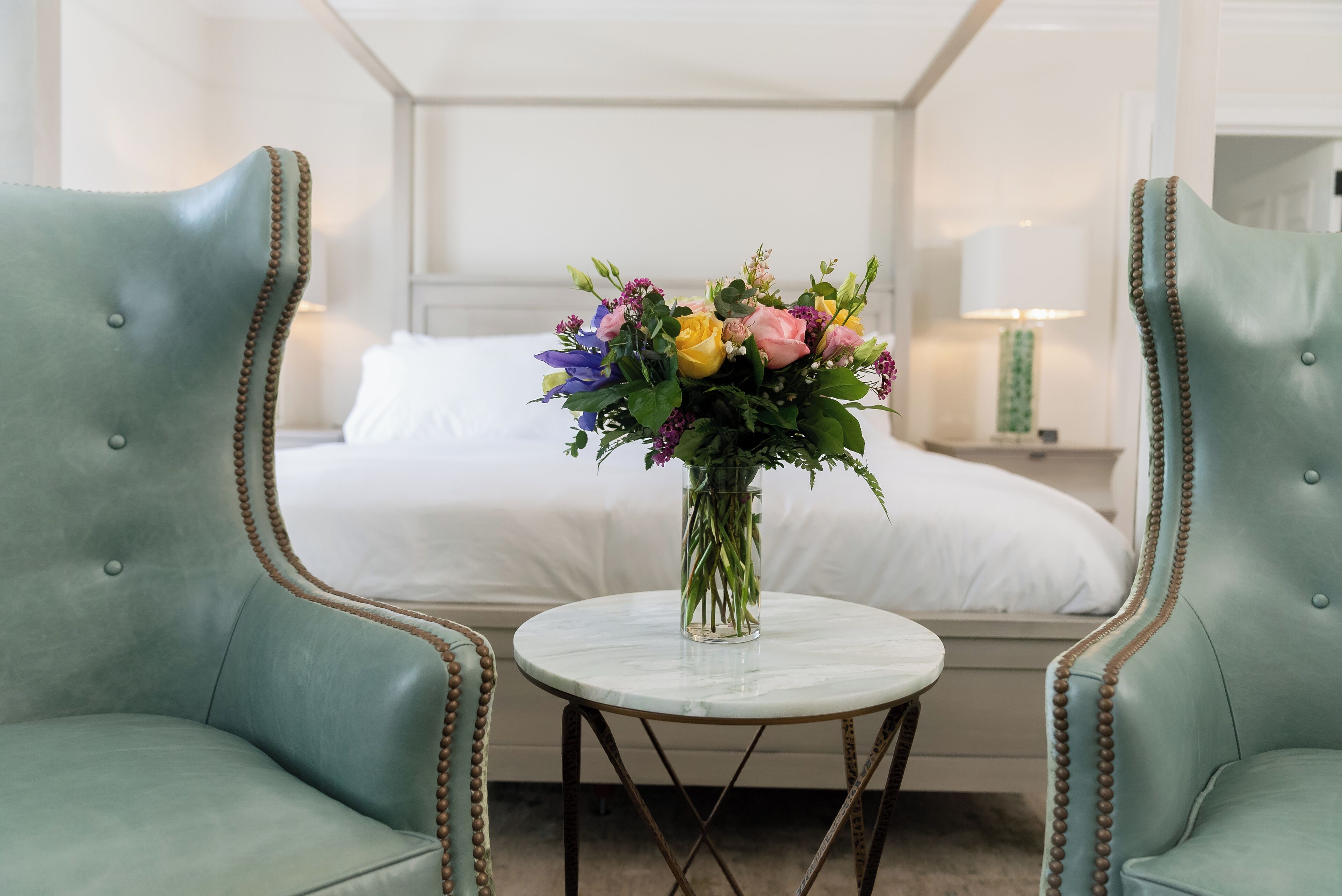 A hotel room with a bed, two green upholstered chairs, a small round table with a vase of flowers, and a nightstand with a lamp.