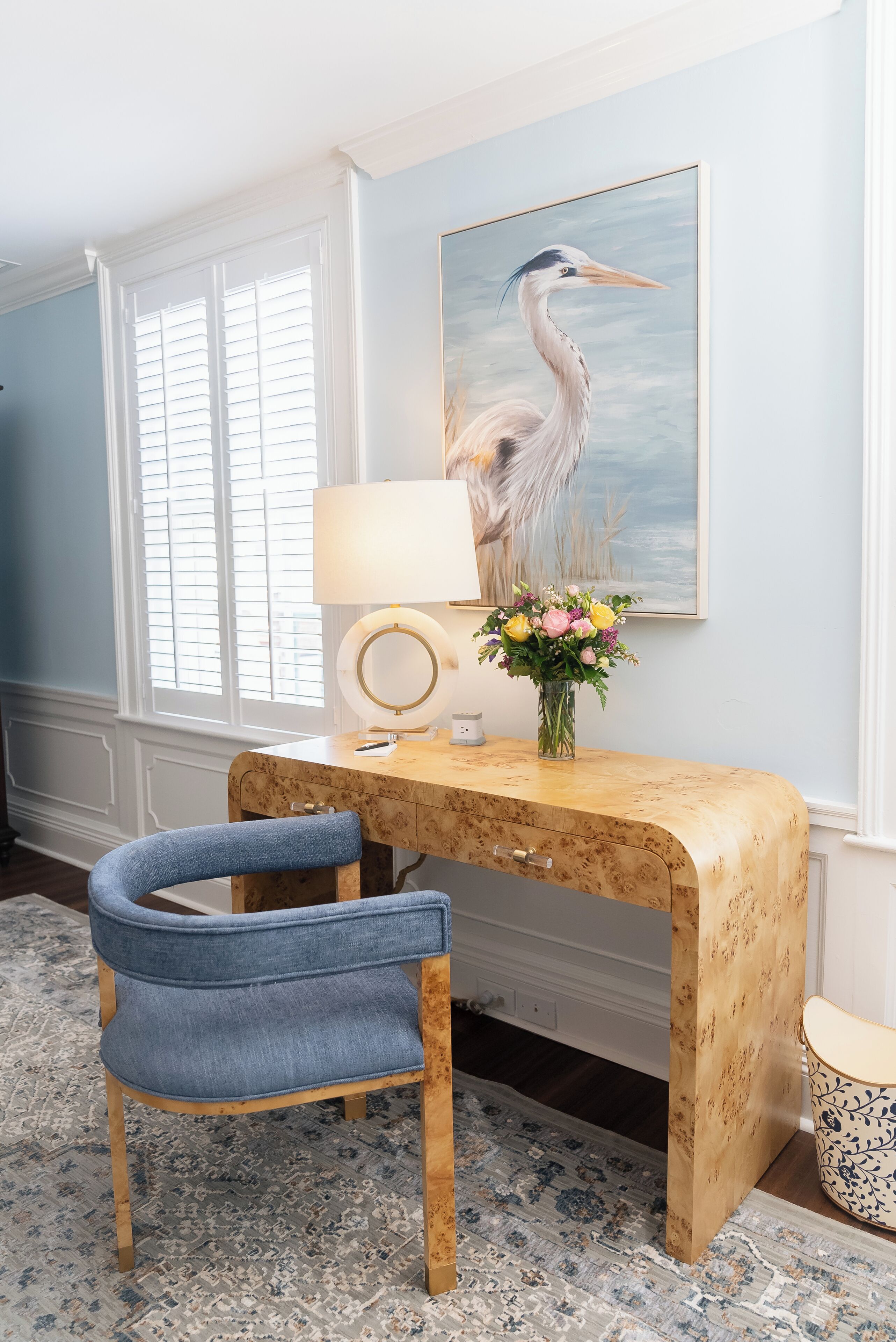 A room with a wooden desk, a blue chair, a lamp, a vase with flowers, and a painting of a bird on the wall.