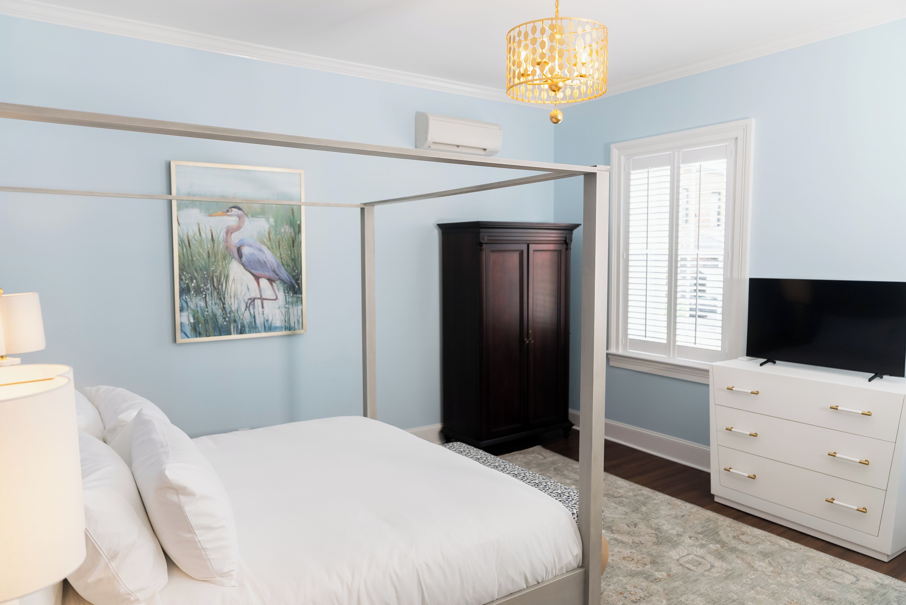 A bedroom with a four-poster bed, a dark wooden cabinet, a white dresser, a flat-screen TV, and a painting of a bird.