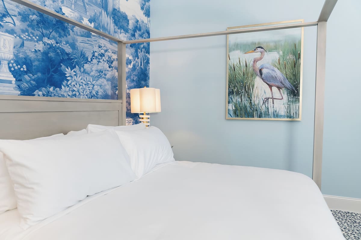 A bedroom with a bed, a nightstand with a lamp, and a framed painting of a bird.