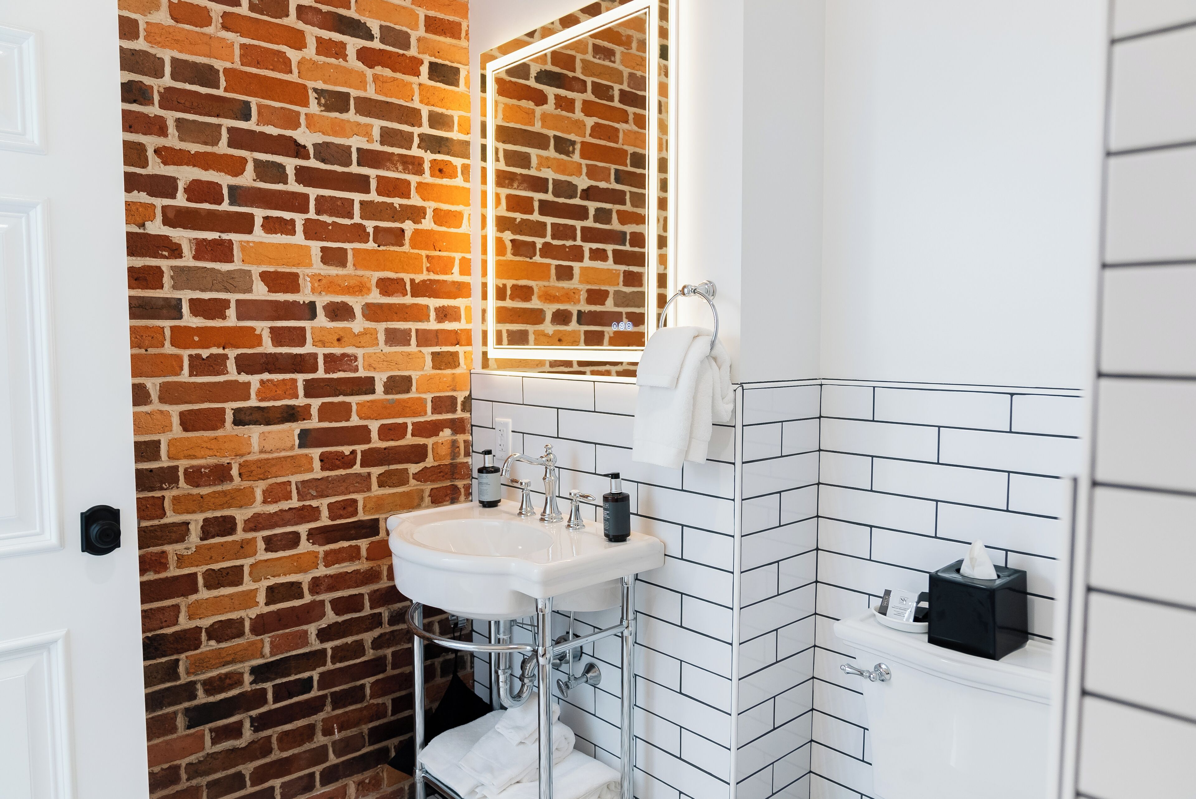 Un baño con pared de ladrillo, salpicadero blanco de azulejos, un lavabo blanco y un espejo.