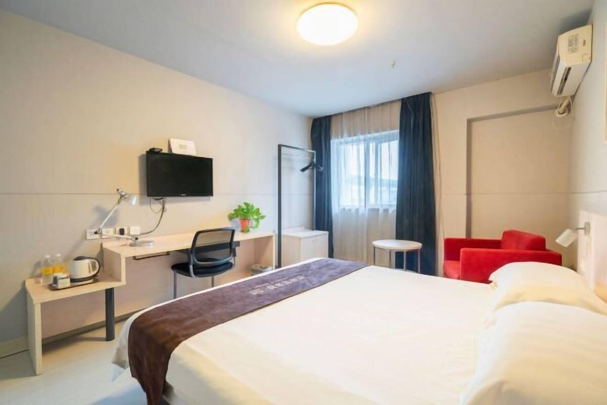 A hotel room with a bed, desk, chair, TV, and a red chair.