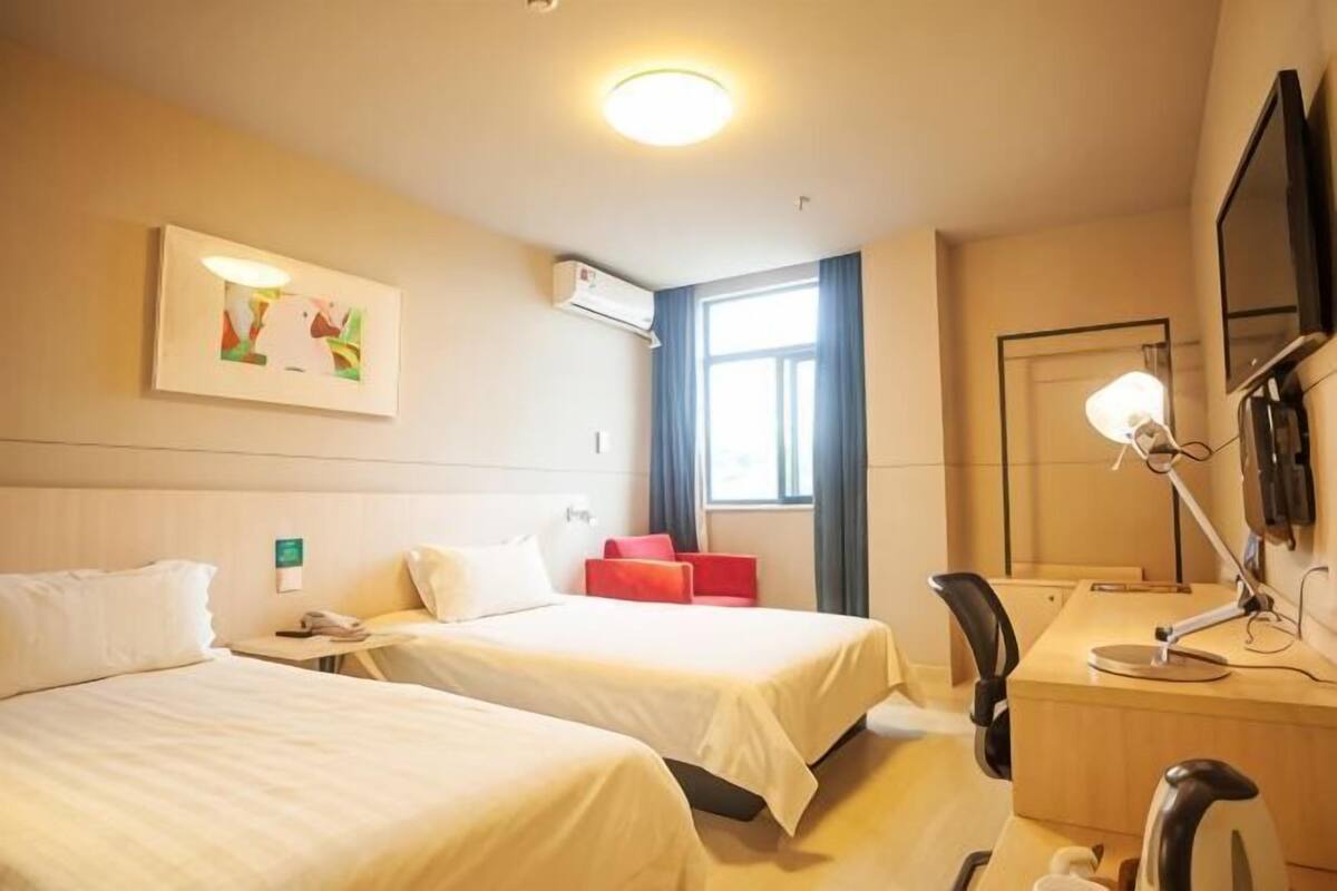 A hotel room with two beds, a desk, a chair, a desk lamp, a mirror, and a window with curtains.