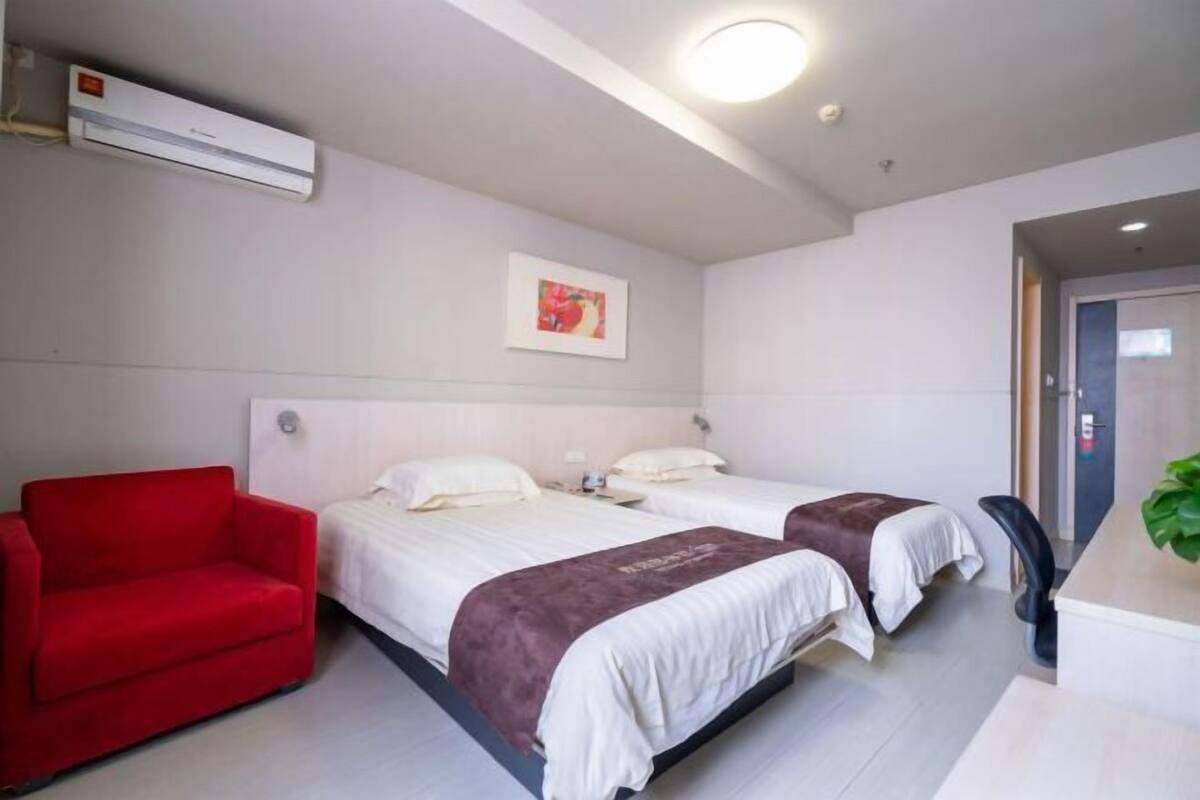 A hotel room with two beds, a red chair, a desk, and an air conditioning unit.