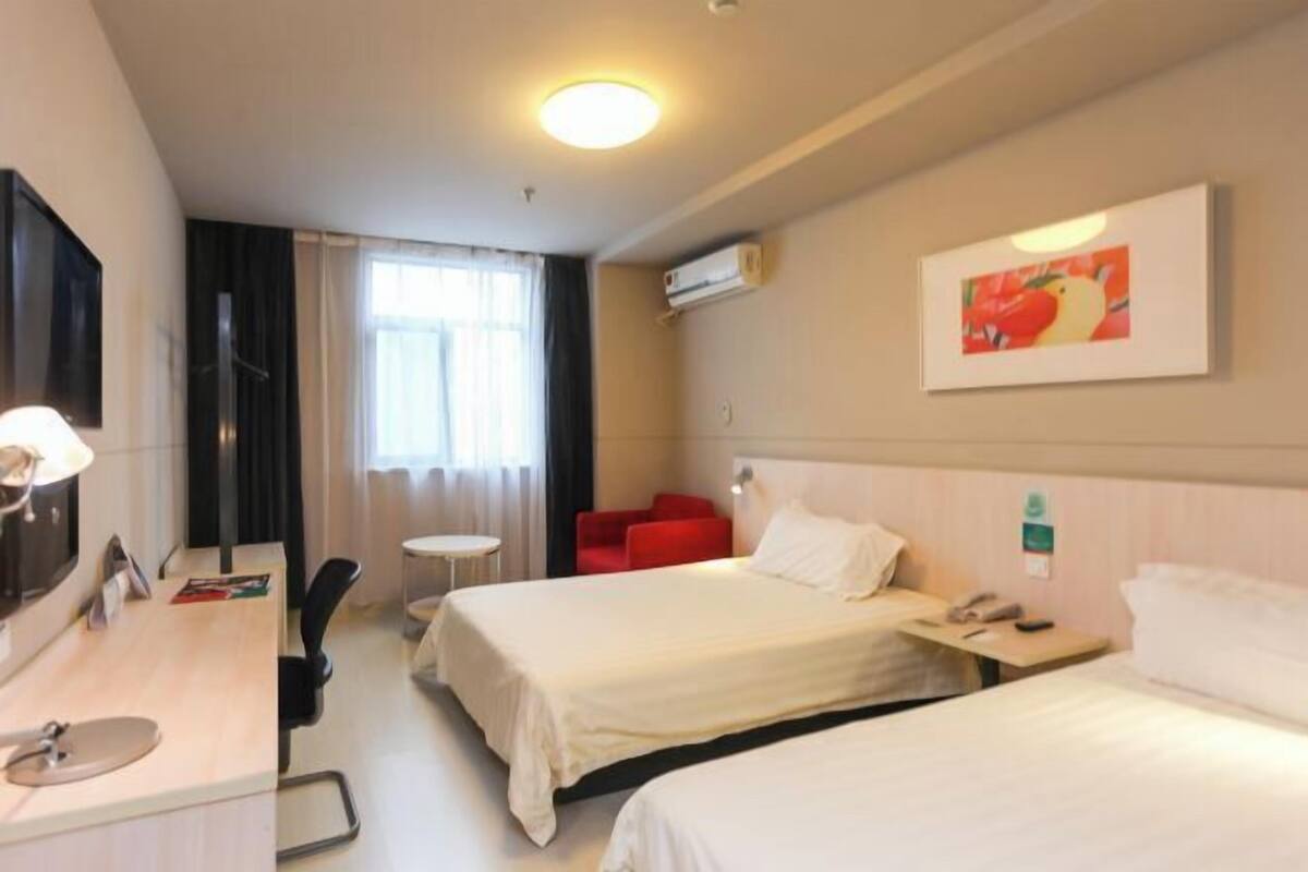 A hotel room with two beds, a desk with a lamp, a small table, a red chair, and a painting on the wall.