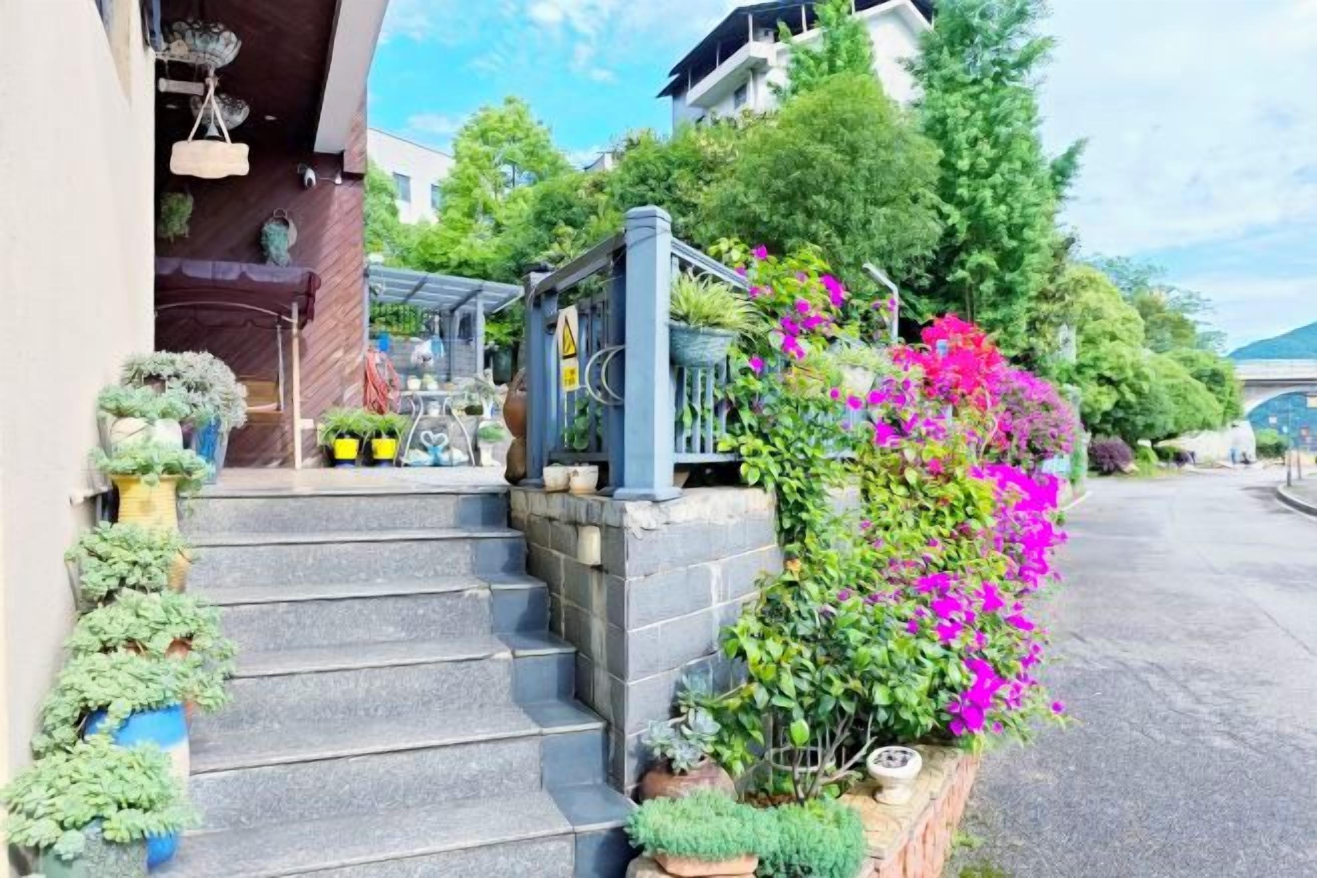 A residential property with a staircase leading to a covered porch, surrounded by potted plants and flowering shrubs.