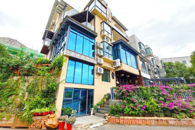 Un edificio moderno de varios pisos con grandes ventanales y balcones, rodeado de abundante vegetación y plantas con flores.