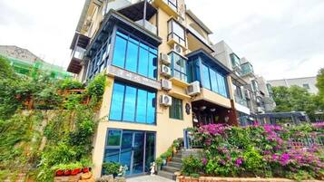Un edificio moderno de varios pisos con grandes ventanales y balcones, rodeado de abundante vegetación y plantas con flores.