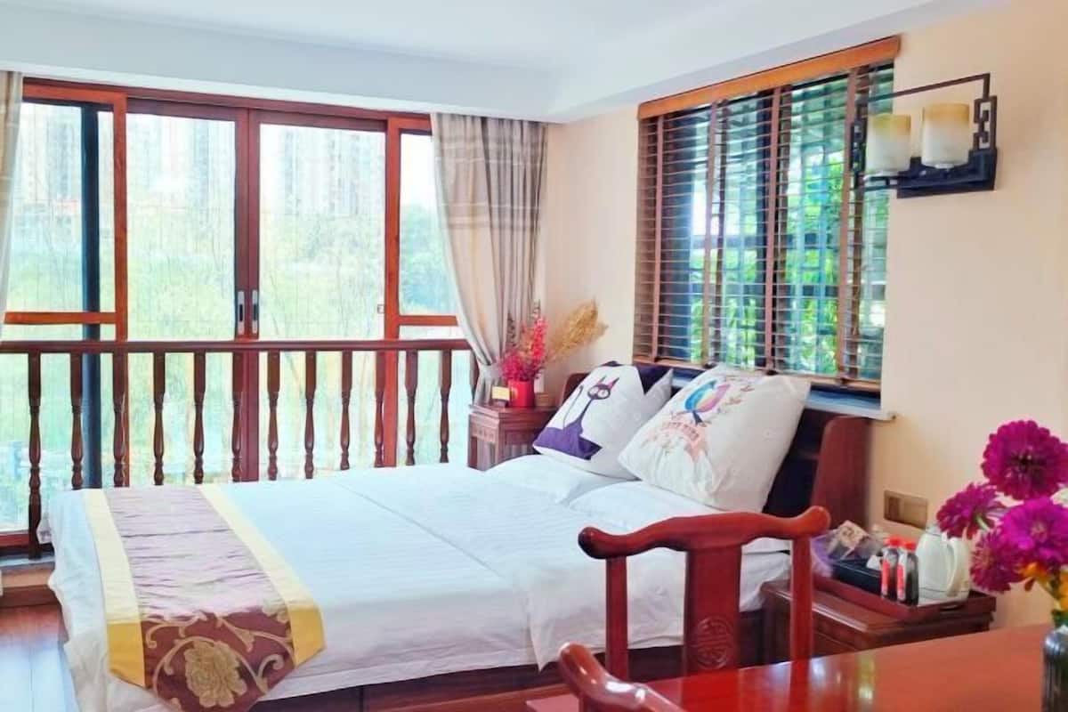 A bedroom with a bed, a wooden chair, a nightstand, a window with blinds, and a balcony with a railing.