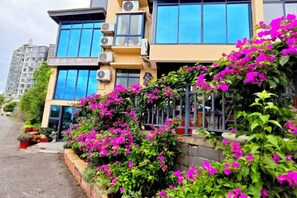 A building with a yellow facade, large windows, and a balcony with pink flowers.