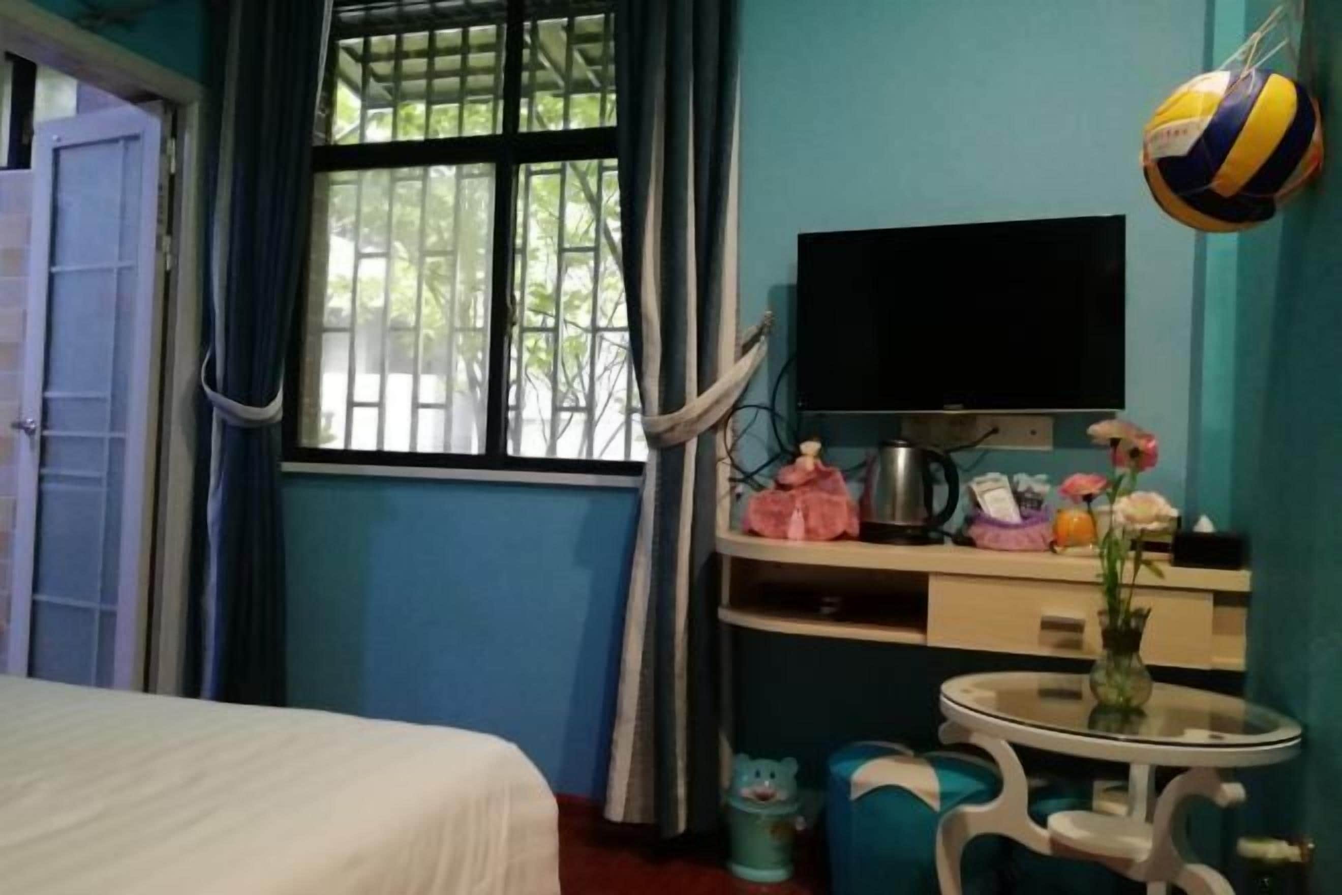 A room with a bed, a TV, a small table with a vase, and a window with curtains.