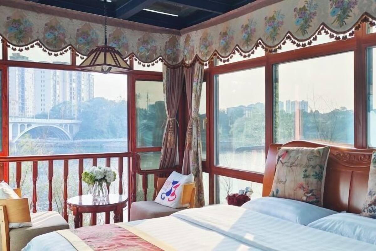 A bedroom with a bed, a nightstand, a chair, a table with flowers, and large windows with a view of a bridge.