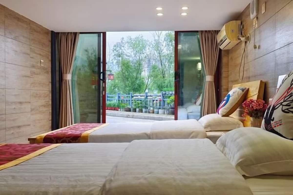A hotel room with two beds, a large window with curtains, a wall-mounted air conditioner, and a view of trees and a bridge outside.