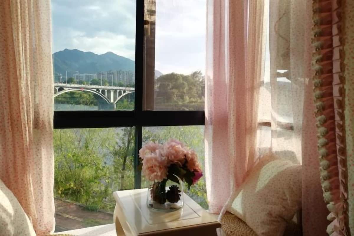 A room with a window, a vase with flowers, and a view of a bridge and mountains.