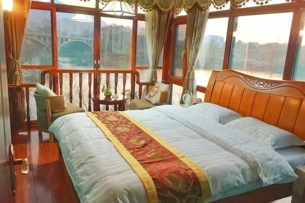 A bedroom with a large bed, a wooden headboard, a small table with a plant, and a view of a bridge through large windows.