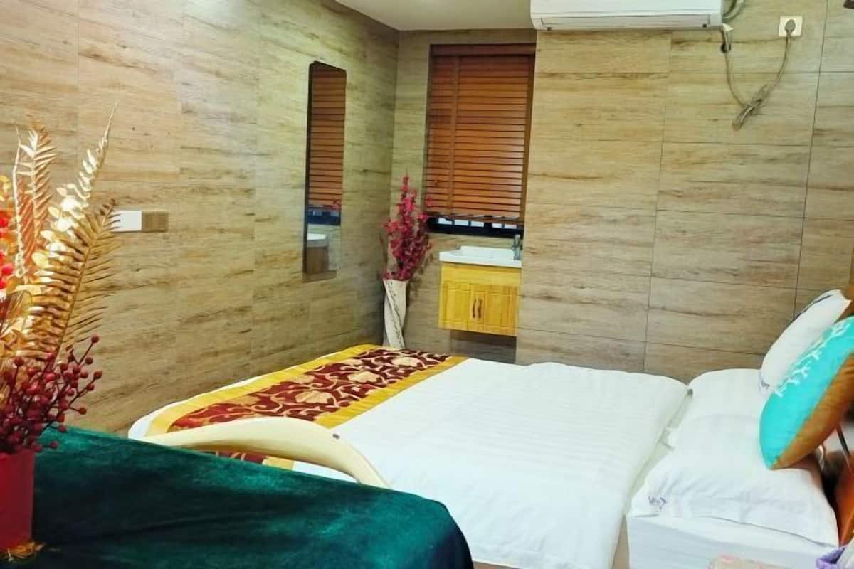 A bedroom with a bed, a small bedside table, a mirror, a window with blinds, and a wall-mounted air conditioner.