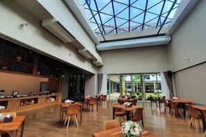 A spacious indoor area with multiple tables and chairs, a high ceiling with a skylight, and a view of greenery outside. - The Cotton Tree Hometel (Khon Kaen)