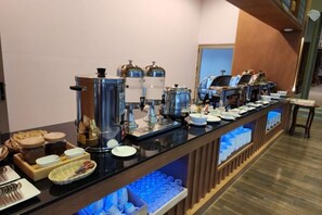Montaje tipo bufé con diferentes dispensadores de comida y bebida, una máquina de café y un mostrador con platos y cubiertos.