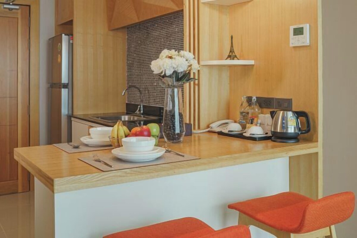 Cocina compacta con encimera de madera, refrigerador, lavamanos y una pequeña zona de comedor con dos sillas rojas.