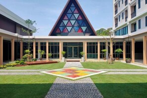 A building with a triangular glass facade, a covered outdoor area with a patterned floor, and a well-maintained lawn. - The Cotton Tree Hometel (Khon Kaen)
