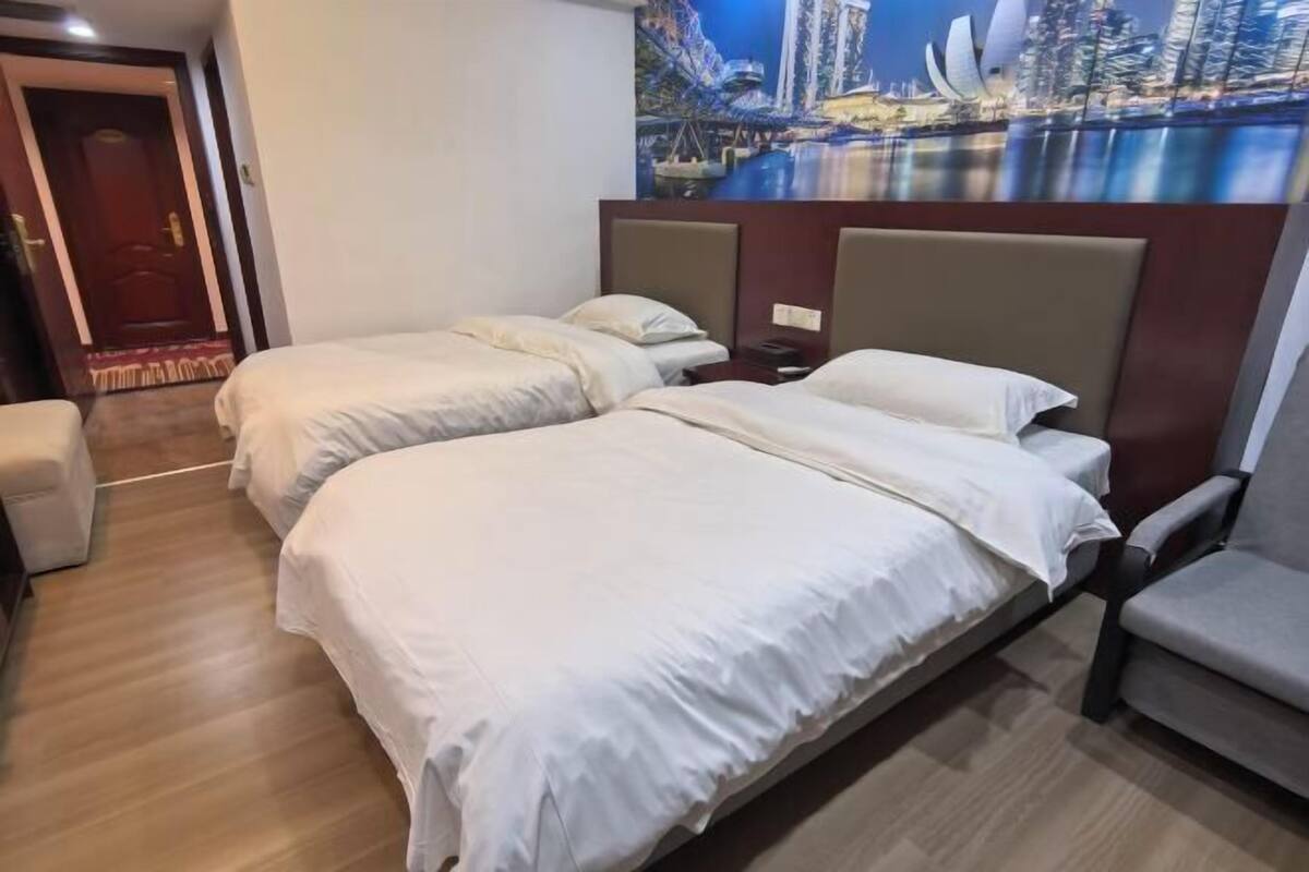 A hotel room with two beds, a chair, a painting of a cityscape, and a door.