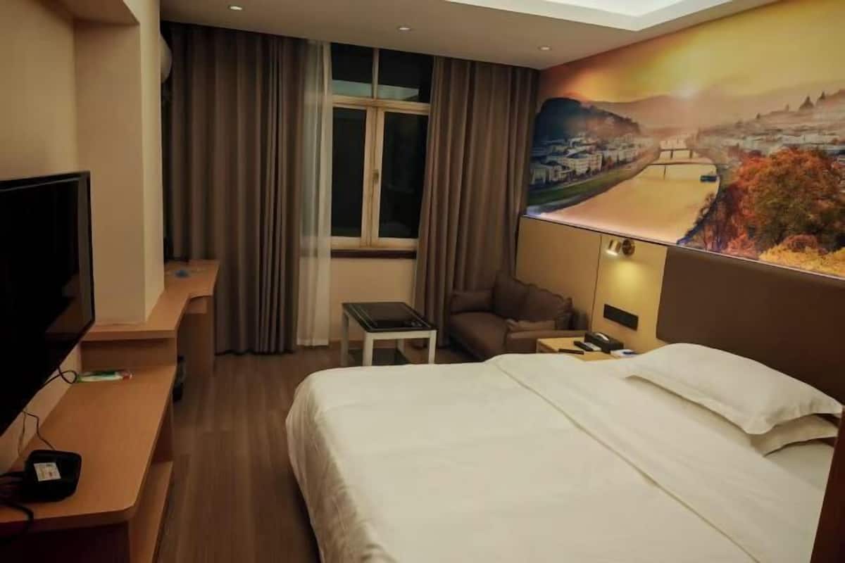 A hotel room with a bed, a desk, a TV, a window with curtains, and a painting on the wall.