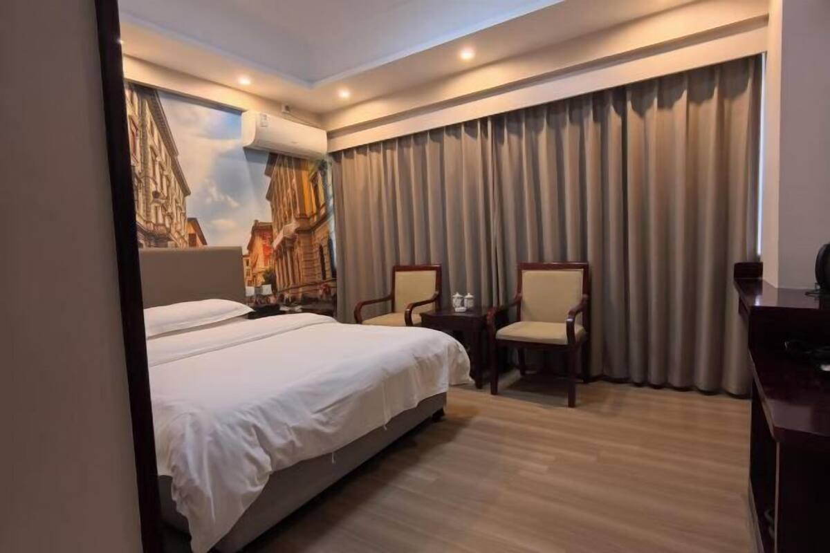 A hotel room with a bed, two chairs, a desk, and a large window with curtains.