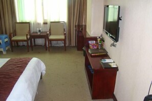 Habitación de hotel con una cama, un escritorio con televisor, dos sillas y una mesita de noche con lámpara.