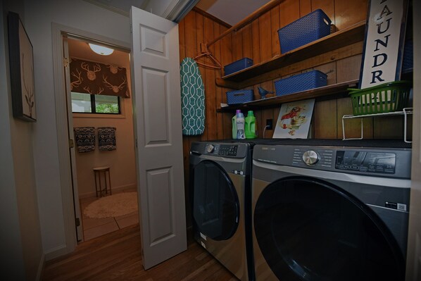 A laundry room with two front-loading washing machines, wooden shelves with laundry baskets, and a wall-mounted ironing board.
