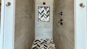A narrow shower area with a chevron-patterned floor, a ceiling-mounted showerhead, and a wall-mounted light fixture. - Sweet Sienna Retreat (Las Cruces)