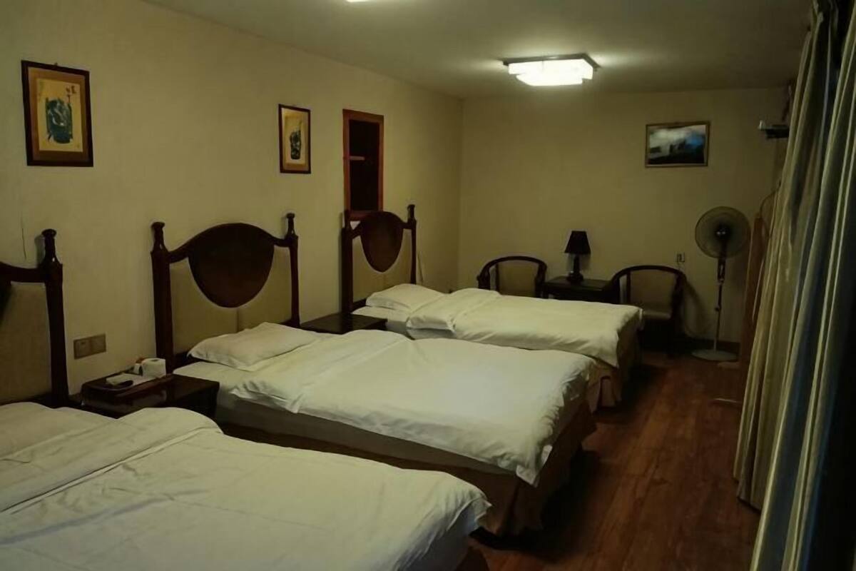 Habitació d'hotel amb quatre llits individuals, una taula de nit amb una làmpada, un ventilador de peu i una finestra petita.