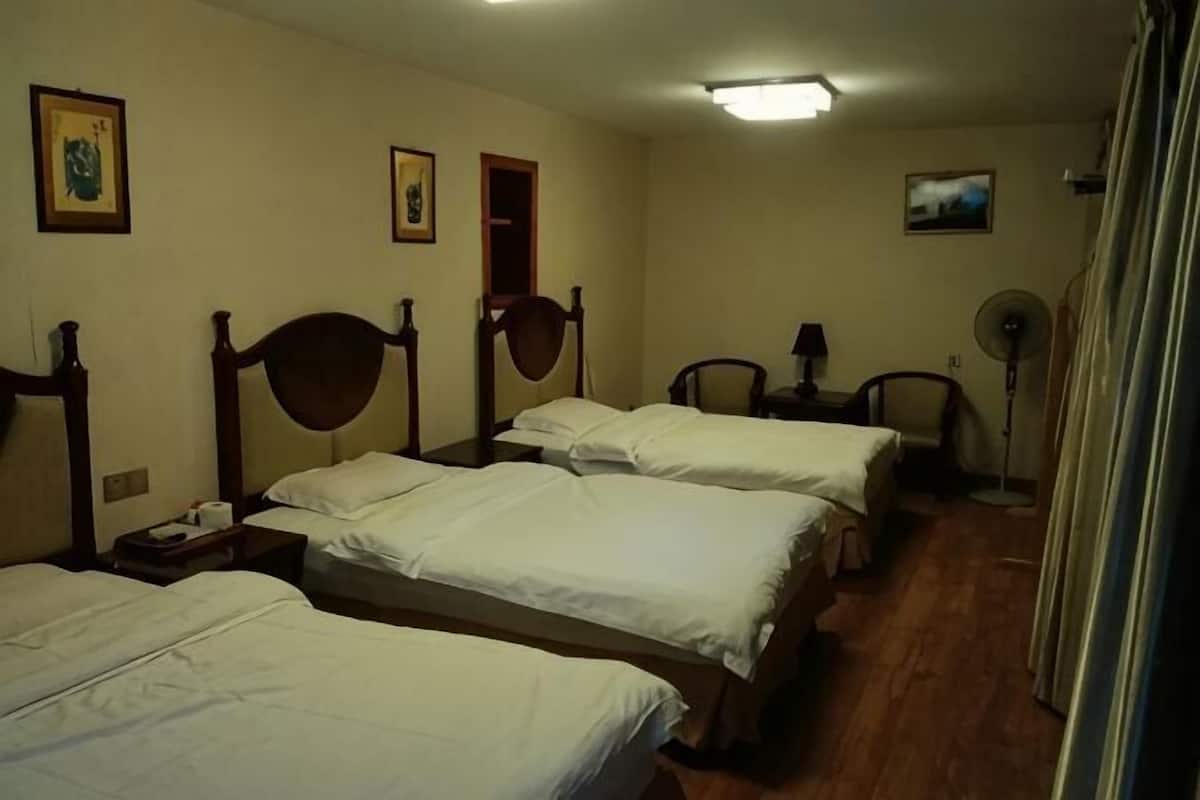 Habitación de hotel con cuatro camas individuales, una mesita de noche con lámpara, un ventilador de pie y una ventana pequeña.